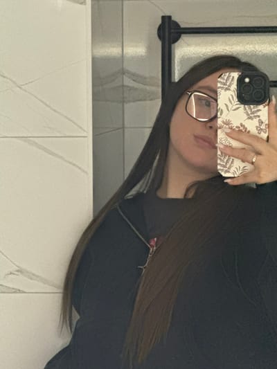 An indoor mirror selfie depicts a female individual in the city of Prikubanskii, Russia. The subject's right profile is visible, partially obscured by a smartphone held in her right hand. She has long, straight, dark hair and wears large, clear-lensed, rectangular-framed glasses. Her right hand displays manicured nails and a ring on the ring finger. The smartphone has a light-colored case adorned with a botanical pattern featuring leaves, flowers, and butterflies, and a triple camera system is discernible. She is dressed in a dark top, possibly a zippered hoodie or jacket, with a necklace visible. The background, reflected in the mirror, shows a tiled wall environment, with white tiles featuring grey veining on the left and darker, patterned tiles with a black towel rack on the right.