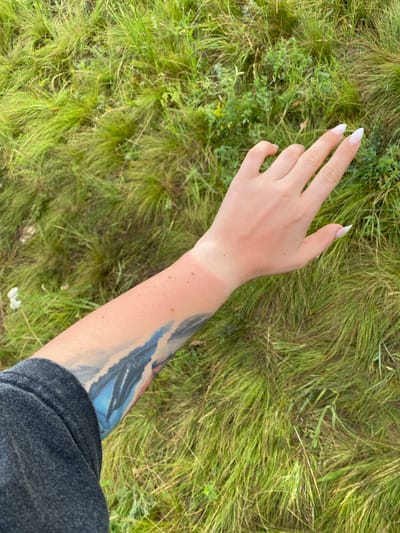 The image shows a person's arm against a background of tall green grass. The arm is tanned on the lower part, indicating a tan line. There is also a tattoo visible on the lower arm, featuring shades of blue and white. The person's hand is raised, with long, white-tipped fingernails. The background suggests an outdoor, natural setting, possibly a field or meadow, with an abundance of lush grass. No specific landmarks or elements are visible to indicate a location within Абзаново, Russia.