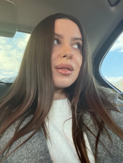 A close-up photograph depicts a female individual seated inside a vehicle, likely in the city of Svinoedovo, Russia. The woman, positioned slightly to the left of the frame, has long, straight, dark brown hair that frames her face and extends over her shoulders. She wears a light-colored, possibly white, knit top beneath a textured grey outer garment, resembling a coat or jacket. Her head is tilted slightly upward, and her eyes, with visible mascara, are directed towards the upper right, away from the camera, conveying a neutral expression. Her full lips are coated with a clear or nude-toned gloss and are slightly parted. The immediate environment suggests a car interior, with a dark headrest or seat back visible behind her left shoulder, a light-colored ceiling panel in the upper left, and a dark car window frame on the right. Through the vehicle's rear and side windows, a bright blue sky with scattered white clouds is visible, and faint outlines of bare trees or light-colored foliage can be discerned in the distant background on the right, indicating an outdoor, daytime setting.