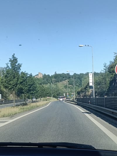 The image shows a view from a vehicle driving on a road in Mladá Boleslav, Czechia. The road is asphalt with white lane markings and a grassy shoulder on the left. A metal guardrail runs along the right side of the road. In the background, there are trees and a hill with ruins of a castle on top. A banner is visible on a pole on the right side of the road, with text that appears to say "GOLF HOTEL". There are several cars and a bus on the road ahead. The sky is clear and blue. The overall scene depicts a scenic drive on a sunny day.