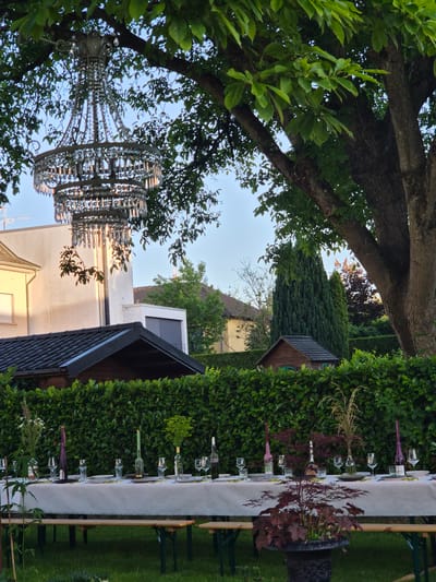 The image depicts an outdoor dining setup in what appears to be a residential garden in Bettembourg, Luxembourg. A long table, covered with a white tablecloth, is set for a meal with plates, glasses, and cutlery. Various decorative items, including bottles, candles, and floral arrangements, adorn the table. A chandelier hangs from a large tree branch above the table, casting a decorative light. The background reveals a neatly trimmed green hedge, a small wooden shed, and parts of houses. The overall scene suggests a peaceful, outdoor gathering or event.