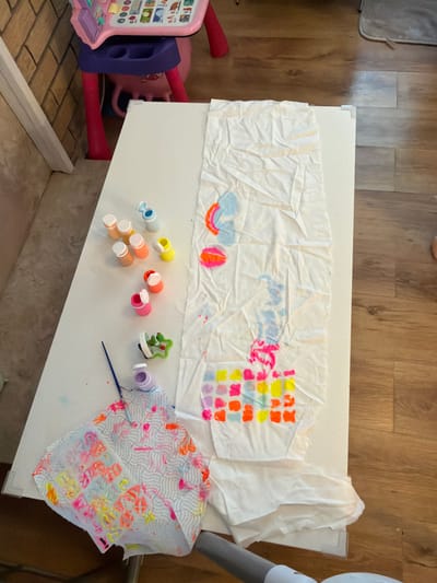 The scene depicts a child's art activity in what appears to be an indoor setting. A white table is covered with a large sheet of crumpled paper, likely used for painting. The paper exhibits various colorful paint splatters, blobs, and abstract patterns. Several small, open containers of paint in shades of blue, yellow, orange, and pink are scattered on the table near the paper, along with a paintbrush. Another piece of crumpled, patterned paper is present towards the bottom right of the table. Part of a pink and purple child's toy sits just to the left of the table. The flooring is a wood-look laminate. The precise location within Bearpark, United Kingdom cannot be determined from the image. No people are visible in the image.