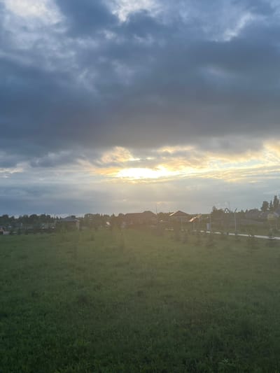 The image depicts a pastoral scene in Kamskiye Polyani, Russia, at dusk. The sky is dramatic, with dark, heavy clouds on the left giving way to streaks of golden light from the setting sun on the right. This transition suggests a clearing of a storm or approaching twilight. Below the sky, a line of houses with distinct roofs, indicative of rural or suburban architecture, is visible, nestled amongst trees. A partially constructed structure, possibly a building or framework, stands prominently near the houses. The foreground is dominated by a lush, green field, suggesting open land or a meadow. The overall impression is one of natural beauty, with the dynamic interplay of light and shadow creating a contemplative and serene atmosphere. The time of day is clearly evening, and the weather is transitioning, likely from rain to clear. There is no visible text in the image.