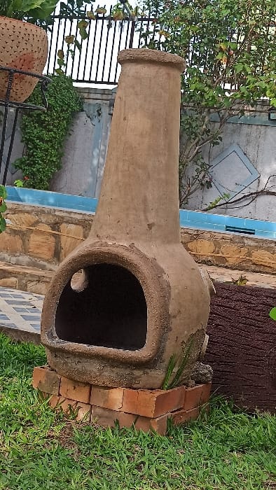 A weathered, light brown earthenware chimenea is centrally positioned on a base of red bricks, surrounded by green grass, in an outdoor setting within Kigali City, Rwanda. The chimenea features a rounded firebox with a prominent arched opening and a tall, tapering chimney stack.
To the left of the chimenea, a dark terracotta pot with perforated patterns rests on a black metal stand, with lush green climbing foliage visible against a light-colored wall and a black vertical-bar metal fence in the background. To the right, a dark brown, textured object resembling a tree trunk or wooden post is partially visible. Further back, a horizontal line of light blue tiles, possibly indicating the edge of a pool or water feature, sits above a section of light brown stone steps and a patterned tiled surface. A light-colored wall with some light blue geometric decorative elements and sparse green foliage constitutes the distant background. No individuals or animals are present. The scene is illuminated by natural light.