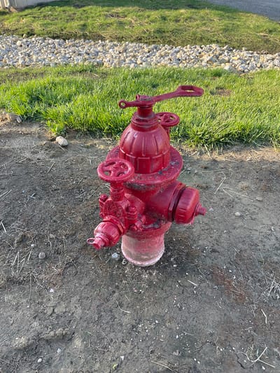 A red fire hydrant stands on a patch of dry dirt and grass. The hydrant has three handles, one large on top and two smaller ones on the sides. It is positioned in front of a strip of gravel that is bordered by green grass. The background is out of focus, but appears to be more grass and a paved surface. The scene is illuminated by sunlight, casting shadows on the ground.