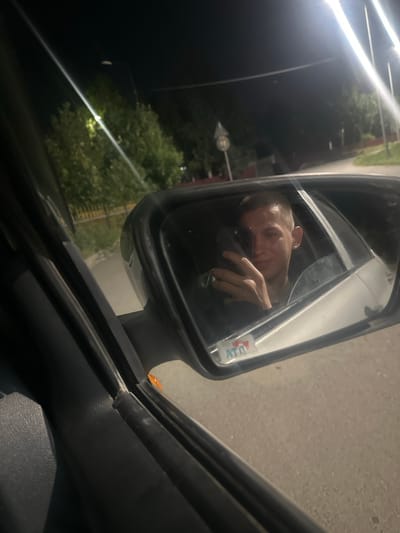 This is a night-time, eye-level, close-up shot taken from inside a car, focusing on the driver's side rearview mirror. The mirror reflects the face of a young Caucasian man with short hair, taking a selfie with his phone. He is wearing a dark t-shirt. The background in the mirror shows a paved road and trees. Outside the mirror, the car's interior trim is visible in the foreground, along with a hint of the exterior through the window, which includes a yellow fence and some foliage. Streetlights are visible in the distance, illuminating the dark environment. The location is identified as Michurino rural district, Kazakhstan.