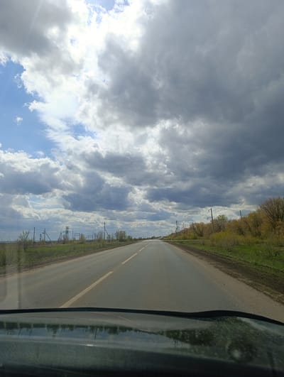 The image captures a first-person perspective from inside a vehicle, looking straight ahead down a two-lane asphalt road. The road is marked with a yellow dashed line down its center, indicating it's a two-way street, and stretches into the distance under a vast, dynamic sky.

On both sides of the road, there's a strip of dirt and then abundant vegetation. The foliage, consisting of bushes and trees, displays varying shades of green and yellowish-green, suggesting a spring or early summer season. On the left side of the road, numerous utility poles with overhead wires run parallel to the road, and a taller, metallic power line tower is visible further in the distance. The right side of the road is bordered by a denser line of trees and bushes.

The sky is a prominent feature, dominated by a mix of heavy, dark grey clouds and patches of bright white clouds. Blue sky is visible in parts, particularly on the left side, where sunlight breaks through the clouds, creating bright spots. The overall impression of the sky is one of changeable weather, possibly before or after a rain shower, or simply a day with dramatic cloud cover.

The reflection of the dashboard and possibly the steering wheel is visible at the bottom of the image, emphasizing the perspective from inside a car. There is no visible text in the image. The scene depicts a rural or semi-rural landscape, consistent with the location context of Лабазы, Russia, likely during daytime.