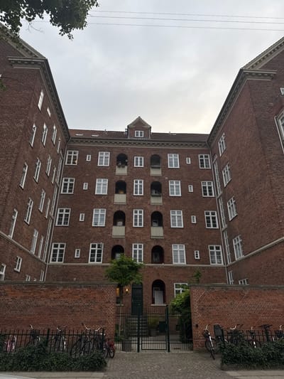This image depicts the exterior of a brick apartment building, likely located in Frederiksberg, Denmark, given its architectural style. The building is U-shaped, with two wings extending forward from a central section. The facade is constructed from red brick, with numerous windows of varying sizes and designs. Some windows are simple rectangular panes, while others feature arched tops or are set within decorative recesses. Several balconies with railings are visible on the central part of the building, some adorned with plants.

In the foreground, a low brick wall runs horizontally across the image, separating the building's courtyard from the street. Behind this wall, a black metal fence encloses a space where several bicycles are parked. The bicycles are of various types and colors. A small patch of green foliage is visible at the base of the fence.

The courtyard area behind the fence contains a paved walkway leading to a dark green door. Steps ascend to this door. A small, leafy tree is situated to the right of the doorway. The sky above is overcast and grayish. Overhead, two thin electrical wires are visible against the sky, stretching across the upper portion of the frame. The overall impression is of an urban residential setting, possibly during daytime given the ambient light.