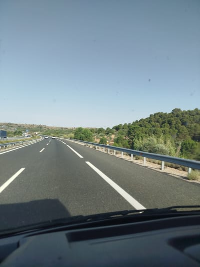 A straight, multi-lane highway stretches into the distance under a clear, bright sky. The road is paved with asphalt and marked with white lane dividers. On the right side of the road, a metal guardrail separates the highway from a gently sloping, tree-covered hillside. The trees appear to be a mix of pines and other evergreen vegetation, with some sparser brush and dry grasses at the base of the hill. In the distance, more rolling hills are visible, also sparsely covered with vegetation. Several cars are driving on the highway, their positions and directions indicating normal traffic flow. A large truck is visible in the distance on an adjacent lane to the left. The view appears to be from the perspective of a driver or passenger inside a vehicle, with the dashboard and windshield wipers visible at the bottom of the frame. The scene suggests a journey through a rural or semi-rural area, possibly in the region of Tébar, Spain, given the arid landscape and vegetation.