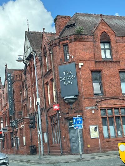 The photograph captures a street scene in Crewe, United Kingdom, featuring brick buildings. At the bottom, the words "ROYAL HOTEL" are displayed. Ascending upwards, there is a set of traffic lights and street paraphernalia such as lampposts. Above this, "The Corner Bar" has a sign, and further up is an "illy" sign. On the right, "PEDLEY STREET" is marked on a sign, along with cycling directions. The sky is partially visible.