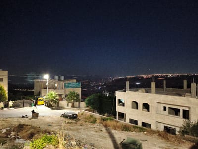 A long shot captures a nighttime view of a hillside settlement in Umm Al-Qatin, Jordan. The dark night sky is dotted with a few stars and some distant city lights. In the foreground, a dirt lot is littered with debris and dry grass. A black car is parked on a paved road in the middle ground. A yellow van is parked near a street light and a large metal gate. Several buildings are visible, including a partially constructed house with exposed rebar. A sign for "Naur First Academy" is posted on one of the buildings. The overall scene is quiet and still, suggesting a peaceful evening.
