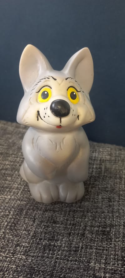 The image shows a small, light gray, plastic toy wolf figurine. The wolf has large, bright yellow eyes with black pupils, a black nose, and a small red mouth. It has black painted eyebrows and a few black dots painted on its cheeks, suggesting whiskers. The figurine is sitting on a textured gray fabric surface, which appears to be upholstery. Behind the figurine, a dark blue background is visible. The lighting suggests an indoor setting. There is no indication of the location in the city of Идрисово, Russia.