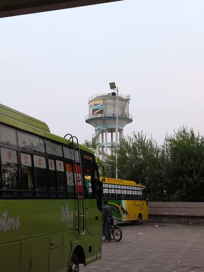 The image depicts an outdoor scene at what appears to be a bus station or depot in Una, India. In the foreground, a bright green bus is partially visible on the left side of the frame, with its side featuring a "MM SINCE 1995" logo. On the right side of the frame, a yellow bus with "Prince" written on its side is parked. A man in a dark shirt and trousers is standing next to a motorcycle in front of the yellow bus. Behind the buses, a large water tower stands tall, with an advertisement visible on its cylindrical section. The ground is paved with interlocking stones. Lush green trees are visible in the background, and the sky is overcast. The scene suggests a typical day at a transportation hub, with vehicles and a few individuals present.