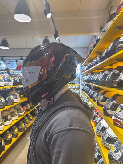 An individual is depicted from the back-right shoulder upwards, positioned inside a retail store specializing in motorcycle gear. The setting is identified as Louis Motorrad Kempten, located at Dieselstraße 9, 87437 Kempten (Allgäu), Germany, based on the distinctive store layout and product offerings.

The subject, of indeterminate gender, is wearing a dark grey zippered hooded sweatshirt over a light-colored neck gaiter and a green undergarment. They are actively trying on a glossy black full-face motorcycle helmet. The helmet features an iridescent, multi-chromatic visor reflecting pink, orange, and yellow hues. A white rectangular product tag, displaying the brand "xNEXX" and a QR code, is affixed to the visor. A chin strap with red and black fastening components is visible beneath the subject's jawline. The helmet also appears to incorporate a small sun visor peak at the top-front.

The surrounding environment consists of multiple brightly lit retail display units. To the right of the subject, extensive rows of bright yellow shelves are densely packed with various full-face motorcycle helmets, each bearing product information tags. Similar shelving units, also stocked with helmets, are visible in the left background, receding into the distance. Overhead, multiple black track lighting fixtures with conical spotlights illuminate the area. In the far-left background, what appear to be racks of shoes or boots are faintly discernible, alongside partially visible red and white balloons. The floor is not visible.
