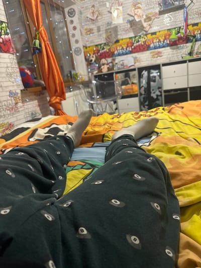 The image presents an indoor scene, specifically a bedroom, located in Matveevskoe, Russia. The perspective is first-person, focusing on the lower body of an individual lying on a bed. The individual wears dark teal pajamas adorned with a white 8-ball pattern and light grey socks. The bed is covered by a multi-colored duvet displaying sections of orange, yellow, and blue.
In the background, a window is visible on the left, draped with an orange curtain tied back. A string of blue-glowing fairy lights is affixed across the windowpane. Adjacent to the window, on the wall, a vertical line of circular smiley face decals is present. The walls are covered with white brick-patterned wallpaper, interrupted by horizontal strips of colorful graffiti-style artwork and several hung or taped pictures/posters. A white radiator is situated beneath the window. A clear acrylic chair stands in the mid-ground, partially obscured. To the right of the chair, a white modular shelving unit contains various objects, including multiple stuffed animals and books. A green desk lamp is visible on the top surface of this unit. A red and blue patterned ball rests on the windowsill.