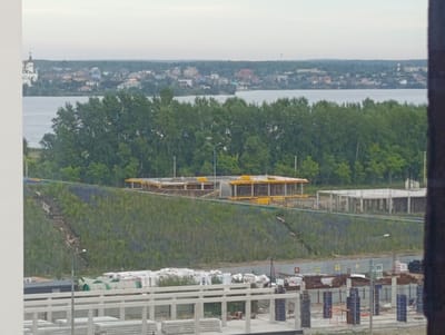 The image shows a construction site in Zapadny, Russia, with a partially built structure in the middle ground and a body of water in the background, bordered by trees and a town. The foreground features stacks of building materials, including white packaged items and cylindrical objects, along with concrete pillars and framing for what appears to be a new structure. A white railing system runs horizontally across the bottom of the image. The middle ground is dominated by a grassy slope with visible construction scaffolding and an unfinished building with a yellow frame. Beyond the slope, a dense line of green trees separates the construction area from a calm body of water. The background reveals a town with various buildings, including a church with a steeple on the left, situated across the water. The sky is overcast.