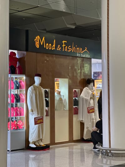 This image captures a retail store display, identified by its signage as "Mood & Fashion By Sud Bio," located in Nouaceur, Morocco. The scene showcases traditional clothing, likely djellabas or similar garments, primarily in white with intricate gold or brown embroidery.

Three mannequins are featured, displaying these outfits. In the foreground, a mannequin on the left wears a white garment adorned with gold trim and a burgundy fez-style cap. Below this mannequin, a glass display case holds an assortment of hats or caps in red and black, some embellished with gold accents. A prominent sign attached to the clothing reads "OFFRE SPÉCIALE JUSQU'À -20%" (Special Offer Up To -20%).

Further into the store, another mannequin is partially visible, dressed in a similar white garment, also bearing a "OFFRE SPÉCIALE JUSQU'À -20%" sign. A third mannequin, visible higher up, appears to be a bust displaying another white garment.

The store's name, "Mood & Fashion By Sud Bio," is displayed in glowing orange text on a dark, geometric-patterned wall panel. The store's interior features wooden paneling and a light-colored, possibly tiled, floor. In the background, a person with dark hair is partially visible, observing the merchandise. The scene suggests a fashion boutique specializing in traditional apparel, currently promoting a discount.