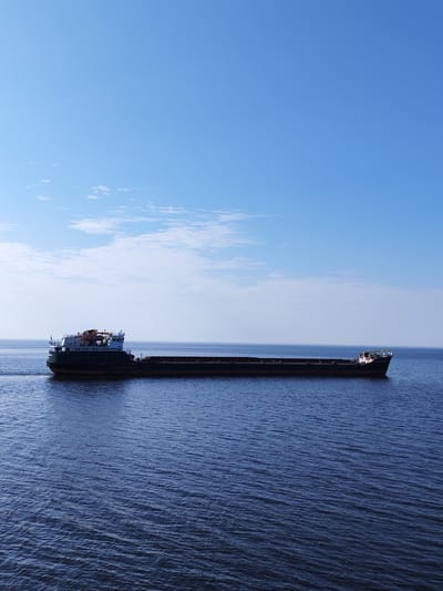 A long, empty barge traverses a body of water under a mostly clear, light blue sky with a few wispy clouds. The water is dark blue and slightly rippled. The barge is dark in color, with a raised section at the front of the vessel. The image does not contain any people. The location, Mologa, Russia, cannot be directly confirmed from visual elements in the image.