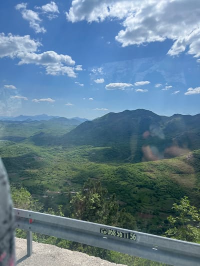The image shows a daytime view of a mountainous landscape with a guardrail in the foreground. The sky is blue with scattered white clouds. The mountain is covered in lush green vegetation. The guardrail is gray and has a sticker with the phone number "SLEP U068 56 11 56" on it. The location is Dobrsko selo, Montenegro.