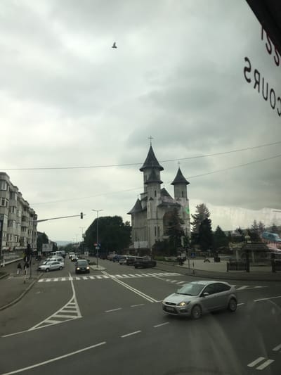 This image depicts an overcast urban street scene in Fălticeni, Romania, looking eastward along Strada Maior Ioan. The photograph appears to be taken from inside a vehicle, evidenced by the reversed text "SS COURS" reflected in the upper right quadrant of the image.
In the background, dominating the scene, stands the Neo-Byzantine style "Sfântul Gheorghe" (Saint George) Church, a prominent landmark in Fălticeni. The church is white with multiple dark, conical roofs topping its two main towers and central dome, each spire culminating in an Orthodox cross. Evergreen trees, likely firs, surround the church on a landscaped area that includes what appears to be a small public square or park with additional decorative structures, including a wooden gazebo-like feature.
In the mid-ground, a multi-lane asphalt road, identified as Strada Maior Ioan, curves through the scene. White lane markings, including a central triangular island with striped chevron markings, delineate traffic flow. A silver second-generation Ford Focus hatchback with Romanian license plates is positioned in the lower mid-ground, driving towards the right. Further down the street, an intersection is visible to the left, marked by a pedestrian crossing (zebra stripes) and a traffic light showing a green signal. Several other vehicles, including a dark minivan, are present on the road and parked along the right side.
To the left of the road, a row of multi-story (approximately 4-5 stories) residential apartment buildings with numerous balconies lines the street, indicative of mid-20th century Eastern European architecture. Two individuals, one adult and one child or smaller person, are walking on the sidewalk adjacent to these buildings near the pedestrian crossing.
The sky is uniformly overcast with light grey clouds. A lone, dark bird is captured in flight in the upper left portion of the sky. In the distant background, faint outlines of hills or mountains are visible beneath the cloudy horizon. Streetlights with modern LED fixtures are positioned along the road.