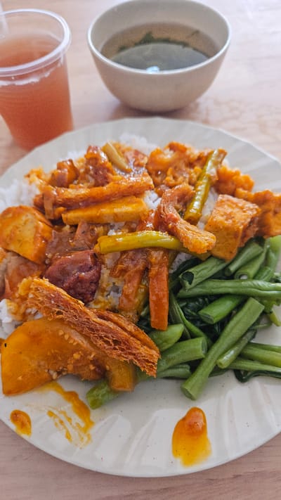 A plate of Vietnamese cơm tấm (broken rice) is presented alongside a cup of soup and a drink. The cơm tấm dish is topped with various fried tofu, beans, and vegetables, covered with a sauce. It is placed on a light-colored wooden surface in a Cam Ranh Ward, Vietnam setting.
