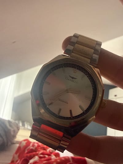 The image shows a close-up of a person's hand holding a gold and silver analog watch. The watch has a light-colored face with gold hour markers and hands. The brand name "GIANELLO" with a winged logo is visible on the dial, along with the word "DIAMOND". The watch band is a combination of gold and silver links. The background is out of focus, but appears to be an indoor setting with diffused lighting. The image is taken at a slight angle, giving a dynamic perspective of the watch. No specific time of day or weather conditions are discernible. The overall impression is one of a personal possession being showcased.