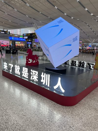 This media file depicts a busy interior scene, likely an airport or train station in Longhua District, China. The primary subjects are people milling about, interspersed with various objects and signage. A large, illuminated cube displaying blue abstract shapes is a central visual element. Adjacent to it, a prominent display features bold white Chinese characters on a black background, which read "深圳北站欢迎您" (Shenzhen North Station Welcomes You) and "来了就是深圳人" (You are a Shenzhen person once you are here). Red signage, possibly indicating departure gates or information points, is also visible.
The setting is a modern, well-lit indoor space with a high, curved ceiling featuring numerous lights. The floor is a speckled gray tile. Several directional signs with Chinese text and arrows guide people, indicating check-in areas, platforms (e.g., "2A-20A", "6A"), and commercial establishments like "全家" (FamilyMart) and "家仕美超市" (Jia Shi Mei Supermarket).
The activity appears to be the normal flow of travelers, with some people standing, others walking, and some seated in waiting areas. The overall atmosphere seems bustling and functional. The time of day is not explicitly stated, but the bright indoor lighting suggests it is daytime. No specific emotions are evident, but the scene conveys a sense of transit and a welcoming message to visitors. The presence of signs in both Chinese and English, along with brand names, points to a modern and international hub.