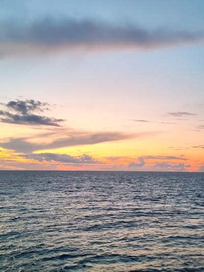 The image shows a serene seascape at sunset. The sky is a gradient of soft blues, pinks, and oranges, with scattered clouds that are illuminated by the setting sun. The clouds range in color from muted grays and blues to vibrant yellows and oranges. Below the sky, a vast expanse of calm, dark blue water stretches to the horizon. The surface of the water is textured with gentle ripples, reflecting the colors of the sky. The scene evokes a sense of tranquility and natural beauty. There are no people, structures, or discernible man-made objects in the image, suggesting a remote or open body of water. The location in the city of Mologa, Russia, while provided, is not visually evident from the image itself, as it depicts a generic seascape.