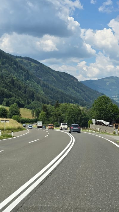 The image shows a two-lane asphalt road curving through a mountainous, forested landscape under a partly cloudy sky. Several vehicles are traveling on the road, including cars, a truck, and a van. The road is marked with white double lines. The surrounding environment consists of lush green mountains covered in trees and some open grassy areas. The sky is a mix of blue and white clouds. The scene appears to be a scenic route in a rural or mountainous region.