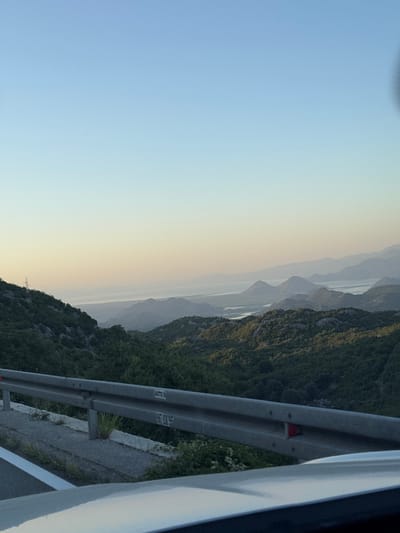 The image depicts a scenic view from a roadside, likely a highway, overlooking a vast valley with rolling hills and distant mountains. The sky is a clear, pale blue, transitioning to a soft, hazy yellow near the horizon, suggesting either early morning or late afternoon. Dense green foliage covers the slopes of the foreground hills, while a network of waterways or a large lake is visible in the midground, surrounded by more layered, misty mountains. A metal guardrail runs along the side of the road in the foreground, separating it from the steep drop into the valley. The road itself is paved with white lane markings. A power line tower is visible on the left side of the frame, nestled amongst the trees. The overall impression is one of natural beauty and tranquility, with the road serving as a vantage point for observing the expansive landscape. The location is specified as Gornja Brova, Montenegro. There are no people or specific actions depicted in the image.