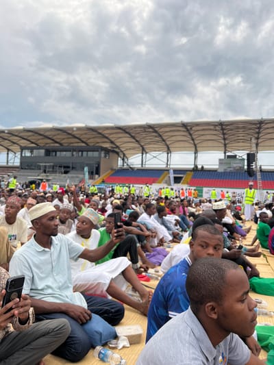 An outdoor scene depicts a large gathering of predominantly male individuals seated on light-colored woven mats on the ground within Amaan Stadium in Zanzibar City, Tanzania. The crowd extends into the midground, with many attendees wearing traditional Islamic headwear (kufis/kofia) and some dressed in white robes or thobes. In the foreground, a man in a light blue collared shirt and a cream-colored kufi is holding a black smartphone horizontally, appearing to record or photograph. To his left, another individual holds a dark smartphone vertically. Plastic water bottles and small food containers are visible on the mats. The stadium structure forms the background, featuring tiered seating in red, blue, and black sections beneath a light-colored, arching canopy roof. Multi-story buildings are integrated into the stadium complex. Elevated platforms with large speakers and a screen displaying "PREMIERE LEAGUE TANZANIA" and "AZIZI" are visible. Several individuals wearing high-visibility yellow vests are present in the background, interspersed within the crowd and near the stadium structures. The sky is overcast with grey clouds.
