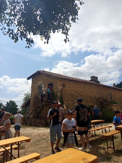 The image depicts an outdoor scene at the Azannes-et-Soumazannes, France historical site on a sunny day. A group of people are gathered around a stone building with a red tile roof. Several individuals are on a wooden staircase leading up to the building's entrance, while others stand on the ground. In the foreground, wooden picnic tables are set up on a dry, grassy area. The sky is blue with scattered clouds, and trees with green leaves are visible in the background, with one prominent tree branch with leaves in the upper left corner.
