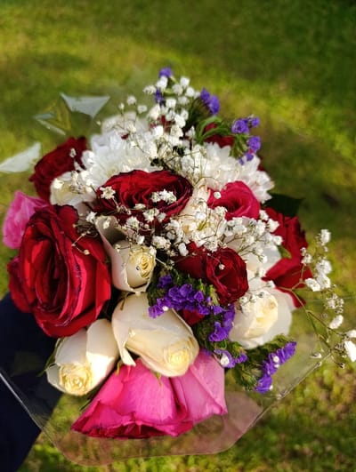 A bouquet of flowers is held in the hand of an unseen person. The bouquet is wrapped in a clear plastic sheet and composed of several red roses, white roses, pink roses, and small white flowers with purple accents. The scene is likely in Kaplelwo, Kenya, as indicated by the provided coordinates.