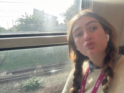 A young adult female is depicted in a self-portrait taken inside a train carriage. She has light brown hair in two braids, light-colored eyes, and poses with a puckered lip expression. White wireless earbuds are in both ears. She wears a cream-colored knitted sweater over a light blue collared shirt. A pink lanyard with "RER E" printed in white text is visible around her neck, indicating the setting is an RER E train. Given the context of RER E, the location is within its service area, which includes Bondy, France.

Through the train window, two railway tracks with sleepers and ballast are visible. Beyond the tracks, green vegetation, including large-leafed plants and bushes, lines the railway. A residential-style building with a prominent sloped roof is discernible in the background under a bright, somewhat hazy sky. Direct sunlight enters from the left, causing a significant glare across the train window. The scene reflects daytime.