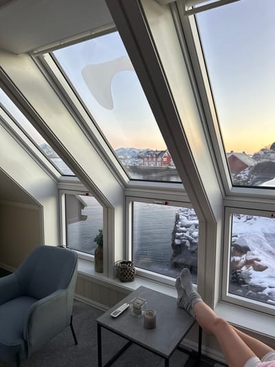 The image depicts an interior space within a building in Stamsund, Norway, showcasing a panoramic view of a winter coastal landscape through multiple white-framed windows. The windows include angled skylights and vertical panes. One upper skylight exhibits a distinct, irregular, axe-head shaped frost or condensation pattern.
Through the windows, a dark body of water, likely a fjord or bay, is visible. To the immediate right, a snow-covered rocky shoreline extends into the water. Across the water, a cluster of buildings is identifiable, with a prominent red house featuring white trim standing out. Snow-capped mountains form the distant background. The sky displays hues of orange and yellow near the horizon, transitioning to light blue overhead, consistent with either sunrise or sunset.
Within the room, the lower legs and feet of a person, wearing light gray ribbed socks, are seen resting on a small, square, gray-topped side table with black metal legs. On this table, a white remote control, a clear glass candle holder with a lit candle, and a second unlit candle are present. To the left, a light gray armchair is partially visible on a gray carpeted floor. A tall vase containing green foliage and a decorative woven lantern are situated on the windowsill beneath the vertical window panes.