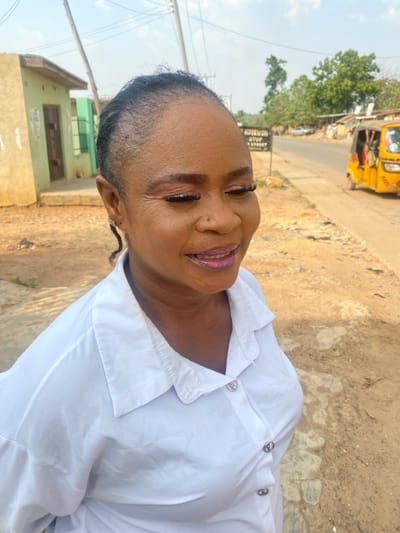 The image captures an adult female in the foreground, situated outdoors on a sunny day in Idiroko, Nigeria. She possesses a medium-dark complexion, with her hair styled in braids pulled back from her forehead. Her face displays makeup, including pink lipstick, false eyelashes, and defined eyebrows, and she exhibits a slight smile with her mouth partially open. She wears a white collared button-up shirt featuring three visible circular, silver-colored buttons. A small, silver-colored earring is visible on her left ear, from which a thin braid extends downwards.
In the mid-ground, to the left, a single-story building with walls painted light green and beige is present, showing multiple doorways. To the right, a dark rectangular sign with white text, partially legible as "AUTOWORLD STOP" and "STOP STREET", stands next to an unpaved, light brown dirt road. Further down the road on the far right, a yellow motorized rickshaw (keke napep) with occupants is visible. The background features sparse green trees and utility poles with overhead lines, all under a clear, light blue sky with minimal cloud cover.