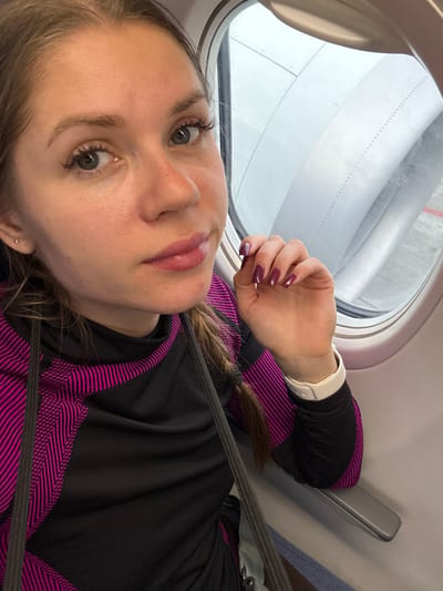 A female individual is depicted in a close-up selfie taken from inside an airplane cabin at an airport in Ürümqi, China.

The subject is a light-skinned female with light brown hair, part of which is visible in a braid over her left shoulder. Her eyes are light in color. She is wearing a black long-sleeved top featuring distinct pink and magenta striped patterns on the shoulders and arms. A grey seatbelt crosses her chest. On her left wrist, she wears a white smartwatch. Her left hand is slightly raised, displaying nails painted a dark burgundy or plum color, with the ring finger's nail appearing a lighter shade of purple. A small, light-colored stud earring is visible in her left earlobe. Her lips are enhanced with a glossy light pink tint.

Through the airplane window to her right, a significant portion of a grey jet engine nacelle is visible. Below the engine, a sliver of tarmac with a red marking line is discernable, indicating the aircraft is on the ground. The exterior sky is overcast and grey. The immediate cabin environment includes a light-colored window frame and a grey armrest.