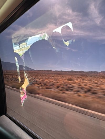 The image shows a view from inside a moving car, looking out the window at a desert landscape. The road is visible in the foreground, running parallel to the car. Beyond the road, the landscape consists of arid, brown terrain with low-lying vegetation. In the distance, there are hills or mountains. The reflection in the car window superimposes the interior of the car onto the landscape. Based on the landscape and the description of "Desert Hot Springs, United States," the scene is located near Desert Hot Springs, and the view is of the surrounding desert.