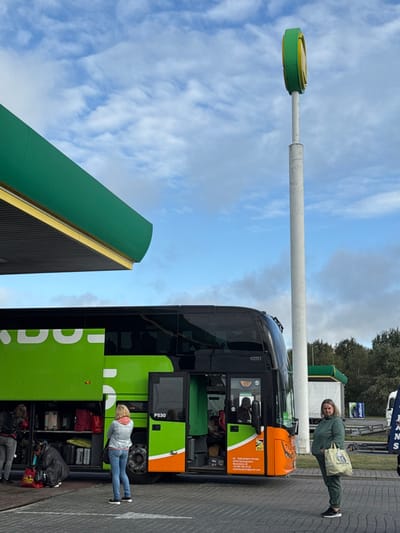 The image depicts an outdoor scene at a service stop in Starovoitove, Ukraine, under a partly cloudy sky. A large, black and green FlixBus double-decker coach, identified by "P530" and displaying "PS TERLETSKY PETRO" with contact information on its lower front panel, is centrally positioned. Its front passenger door is open, revealing an individual seated inside. A side luggage compartment door is also open, exposing multiple suitcases, bags, and red parcels. Two individuals are located near this compartment; one kneels, actively handling luggage, while another stands next to them. A third woman, with blonde hair and wearing a light blue denim jacket over a pink top and blue jeans, stands facing the bus, observing the luggage area. To the right of the bus, a fourth woman, dressed in a matching olive-green tracksuit, stands facing the viewer, holding a light-colored canvas bag with blue lettering in her left hand. The immediate environment includes a green overhead canopy covering part of the scene and a tall white pole topped with a green and yellow circular sign. The ground is paved, and trees are visible in the distant background.