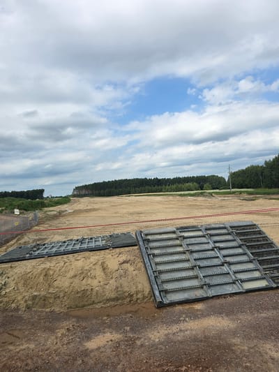 This is a landscape photograph taken on a cloudy day in Omulno, Poland. The foreground features a construction site with piles of sand and construction materials. Two large metal grates are laid out on the sand, likely for temporary road surfaces or ground support. A red caution tape is strung across the scene, indicating a restricted area. Beyond the construction site, there is a large expanse of cleared land, possibly for future development. In the distance, a dense line of trees forms a natural boundary, with a few scattered utility poles visible. The sky is filled with dramatic clouds, a mix of white and gray, with patches of blue sky peeking through. The overall impression is one of an ongoing construction project in a rural or semi-rural setting.