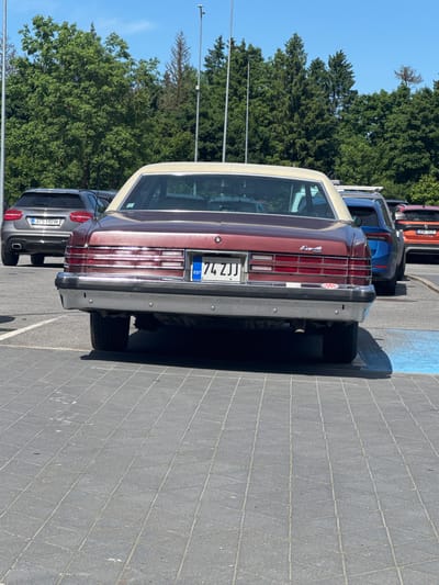 In the city of Tabasalu, Estonia, on a sunny day, a row of parked cars is visible on an outdoor paved lot. The foreground is dominated by the rear of a maroon 1974 Pontiac Grand Ville sedan with a beige vinyl roof. The car has a chrome bumper and a license plate reading "74 ZJJ EST". To the left of the Pontiac is a silver Mercedes-Benz SUV with the license plate "375 HBW". Behind the Pontiac, other vehicles are partially visible, including a blue SUV and an orange car. The background features a dense line of green trees under a clear blue sky.