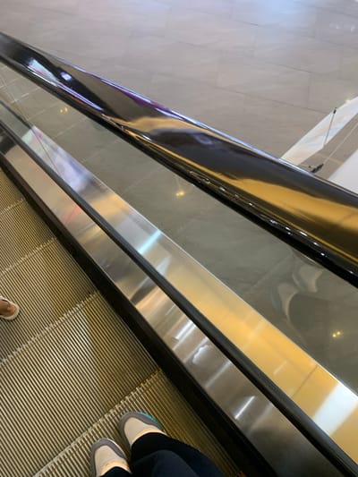 The image captures an overhead perspective looking down an escalator inside a modern building, likely a mall or commercial complex in Samarinda, Indonesia. The escalator steps, visible in the foreground and extending upwards, are a metallic, ribbed surface with a golden or bronze hue. On these steps, the feet of at least one person are visible, wearing light-colored sneakers with dark pants. Another foot is barely visible at the bottom left.

The escalator's handrail, located on the left side of the frame, is sleek, black, and highly reflective, showing distorted reflections of interior lighting and possibly parts of the ceiling or upper floors. Adjacent to the handrail are polished metal panels that also reflect light and elements of the surrounding structure, creating bright streaks and abstract shapes. Beyond the escalator, a light-colored tiled floor with a geometric pattern is visible, suggesting an open floor plan in the building. The overall scene indicates an indoor setting, illuminated by artificial lighting, with a sense of movement as the escalator ascends or descends. No discernible text or specific emotions are evident.
