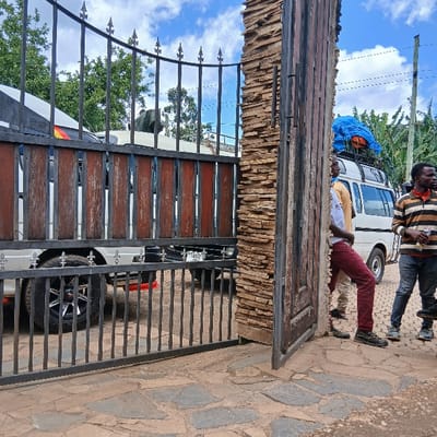 The scene depicts an outdoor gate entrance in Mweka, Tanzania. The gate consists of dark metalwork with vertical bars and decorative finials, featuring wooden panels on its left, mostly closed section. The right section of the gate is partially open, revealing a substantial pillar clad with stacked natural stone. The ground in the foreground is paved with irregularly shaped flat stones.

Behind the gate, two vehicles are present. A white pickup truck or utility vehicle, equipped with a white canopy, is partially visible through the closed section of the gate. A dark-furred animal, consistent with a goat, is situated on the rear bed of this truck. The vehicle's license plate "T 868" is partially discernible. Further to the right and behind the open gate, a white passenger van, possibly a minibus, is visible with luggage covered by a blue tarp secured to its roof rack.

Three adult males are standing to the right of the partially open gate. The leftmost male is partially obscured, wearing a light-colored shirt and dark trousers. The central male wears a light-colored shirt, red trousers, and dark shoes, leaning against the stone-clad gate pillar. The rightmost male wears a long-sleeved horizontally striped shirt (brown, orange, white), dark jeans, and dark shoes, holding a clear plastic bottle in his left hand. All three individuals are looking towards the left, off-camera. The background shows green foliage from trees and bushes under a bright blue sky with scattered white clouds. The scene is illuminated by direct daylight.