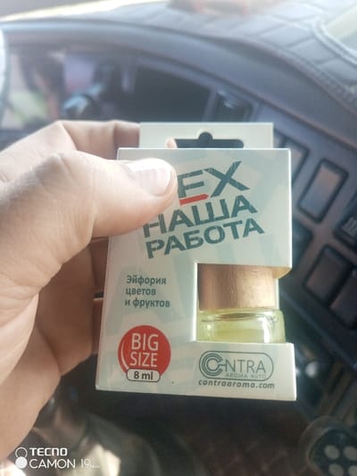 A human hand holds a white cardboard package containing a small glass bottle filled with a light yellow liquid, sealed with a wooden stopper. The package's front displays text in Russian, including "EX НАША РАБОТА" (EX OUR WORK), "Эйфория цветов и фруктов" (Euphoria of flowers and fruits), "BIG SIZE 8 ml", and the brand "CONTRA AROMA AUTO" with its website "contraaroma.com". The hand's thumb partially obscures the upper portion of the text. The background is a blurred view of a vehicle's interior, showing a dark, textured dashboard with integrated controls and a light source indicating an exterior environment. The specific location within the city of Pridorozhnyi, Russia, is not ascertainable from the visual evidence.