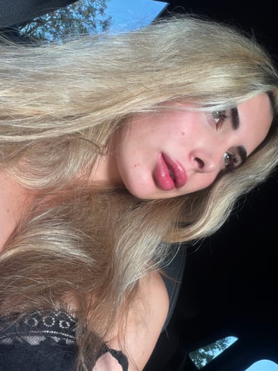 The image shows a close-up, angled portrait of a young woman with long, blonde hair and full lips. She is wearing a black lace top. The background is dark, with a sliver of a blue sky and green foliage visible through what appears to be a car's sunroof. The lighting suggests it is daytime. There is no indication of the city of Sant Vicent del Raspeig, Spain in the image.