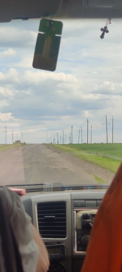 The image captures a sideways view from inside a vehicle, looking out onto a rural landscape in Shelestove, Ukraine. The scene is dominated by a long dirt or gravel road stretching into the distance, bordered on one side by a lush green field, likely agricultural. A continuous line of wooden utility poles with overhead power lines runs alongside the road, extending towards the horizon under a bright sky with scattered white clouds. A faint white contrail is visible in the sky.
Inside the vehicle, elements of the interior are visible. In the upper left, a dark, ornate cross hangs from a chain, possibly from the rearview mirror. Below it, a blurry, rectangular object with a green tint, possibly a phone or a pass, is seen. On the right side, parts of the vehicle's dashboard are visible, including a silver-colored air vent with vertical slats and a small, unreadable digital display unit. An orange piece of fabric or trim is in the upper right corner. In the lower right, a person's forearm and hand rest inside the vehicle, with a white, soft object (like a cloth or tissue) nearby.
The image portrays a daytime scene, suggesting fair weather conditions during a journey or travel through the countryside. There is no discernible legible text within the frame.