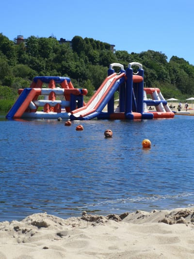 In Yantarny, Russia, a sunny day showcases a large, inflatable water park situated in a body of water adjacent to a sandy beach. The water park is a vibrant structure of red, white, and blue inflatable elements, featuring slides and climbing areas. Several people are visible in the background, some standing under white umbrellas on the beach, while others are near the water park. In the foreground, the sandy beach meets the blue water, with three orange buoys scattered in the water in front of the inflatable structure. Trees and buildings line the horizon behind the water park. The sky is clear and blue.