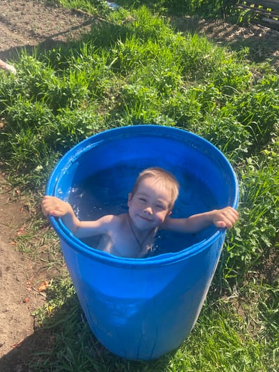A young, fair-skinned child with short blonde hair is submerged in a blue plastic barrel filled with water. The child, appearing to be between the ages of 3 and 6, is positioned inside the barrel such that only their head, shoulders, and arms are visible above the water's surface. They are smiling directly at the camera and grasping the rim of the barrel with both hands.

The barrel is situated outdoors, adjacent to a patch of dense green grass. The ground surrounding the grass area is mostly bare and appears to be dirt or soil. The lighting is bright, suggesting the photograph was taken on a sunny day. Part of a wooden fence is visible in the upper left corner. Based on the visual content of the image, there is no verifiable information about the specific location of Узяр, Russia.