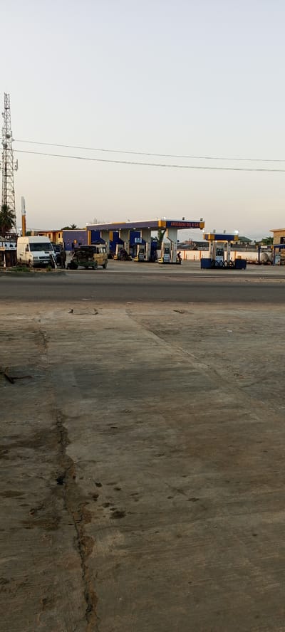 This media file depicts an active petroleum filling station identified as "ARIGBABU OIL & GAS" in Alasia, Nigeria. The scene is bathed in soft, diffused light, suggesting either early morning or late afternoon/early evening under a clear, pale sky.
In the foreground, a large, worn concrete or paved area dominates, showing numerous cracks and dark oil stains. The main subject, the gas station, is positioned in the mid-distance. It features distinct blue and yellow canopies sheltering multiple fuel pumps. Several vehicles are visible, including a white van parked on the far left and an older van with its hood open, indicating it might be undergoing repair or inspection, with an individual attending to it. Other vehicles are seen at the pumps, alongside several people who appear to be either refueling their vehicles or working at the station. A person on a motorcycle is also present near the pumps.
On the far left, a tall, metallic communications or cell tower stands prominently against the sky. Power lines stretch horizontally across the upper portion of the image. Behind the main gas station structures, storage tanks are visible on the right, and other buildings can be faintly discerned in the distance, providing a backdrop to the bustling but routine activity at the filling station.