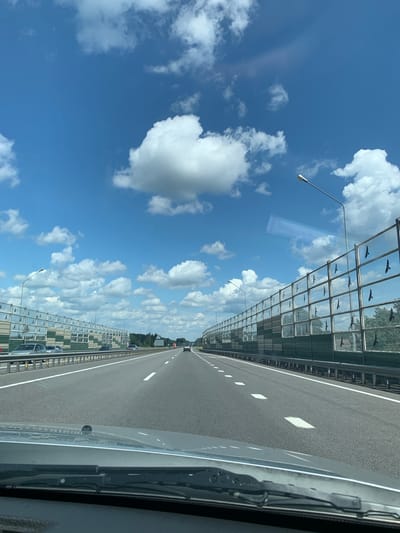 The image shows a forward-facing view from inside a vehicle traveling on a multi-lane highway. The highway is flanked by tall sound barriers with reflective panels that have bird silhouette markings. The sky is bright blue with scattered white cumulus clouds. Streetlights are visible along the highway. Traffic consists of several cars and a truck in the distance. The environment appears to be rural or suburban, with trees visible beyond the sound barriers. The condition of the road is good, with clear lane markings. The foreground shows the dashboard and windshield wipers of the vehicle.